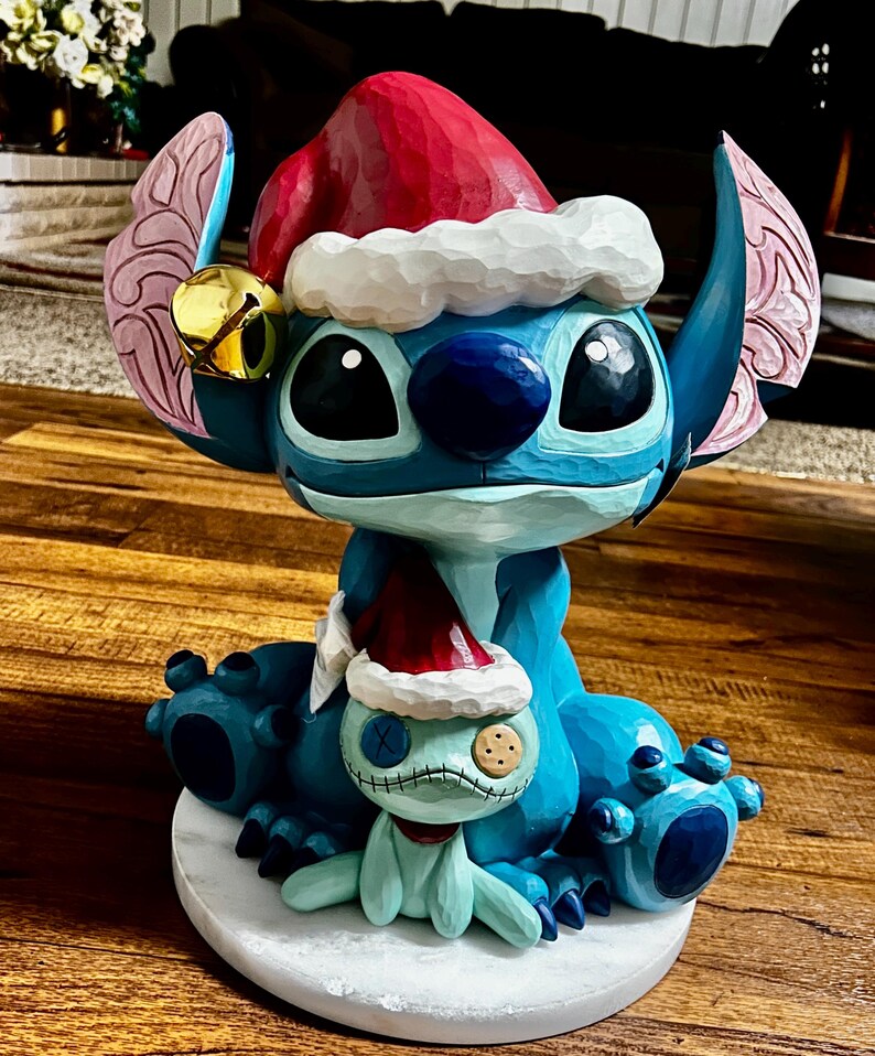 May include: A blue cartoon character figurine wearing a red Santa hat with white trim and a gold bell. The figurine is holding a smaller version of itself, also wearing a Santa hat. The figurine is sitting on a white marble base.