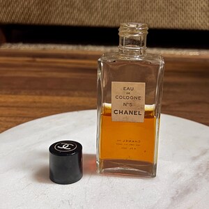May include: A rectangular glass bottle of Chanel No. 5 Eau de Cologne with a black cap. The bottle contains a golden-colored liquid and has a label with the brand name and product details. The cap is off to the side.