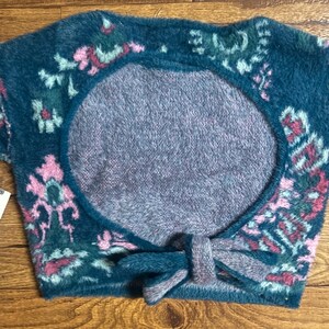 May include: A teal blue fuzzy sweater with a pink and green floral pattern. The sweater has a large open back with a tie detail.