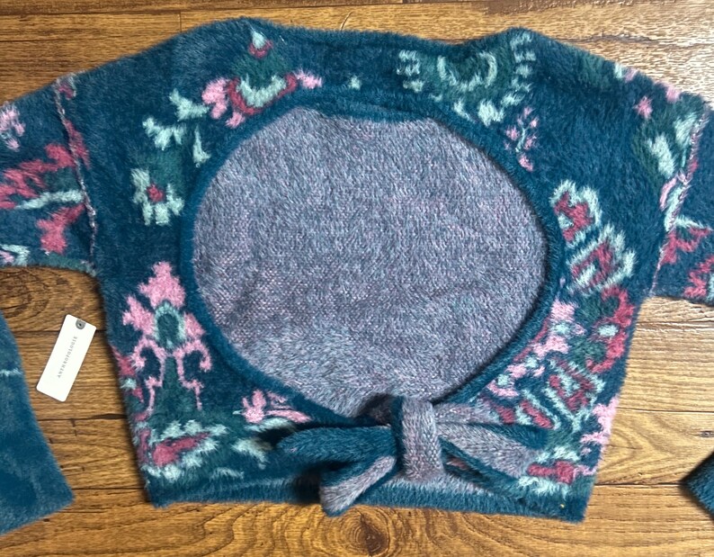 May include: A teal blue fuzzy sweater with a pink and green floral pattern. The sweater has a large open back with a tie detail.