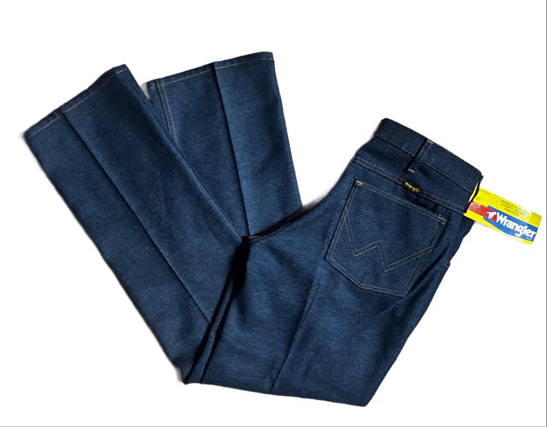 Może przedstawiać: Ciemnoniebieskie dżinsy Wrangler o klasycznym kroju. Dżinsy mają tylną kieszeń z logo Wrangler oraz ż&oacute;łto-czerwoną metkę. Dżinsy są złożone i prezentowane na białym tle.