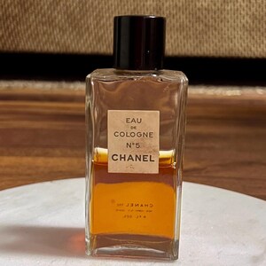 May include: A rectangular glass bottle of Chanel No. 5 Eau de Cologne. The bottle has a black cap and a beige label with black text. The liquid inside is amber-colored. The bottle is sitting on a white surface.