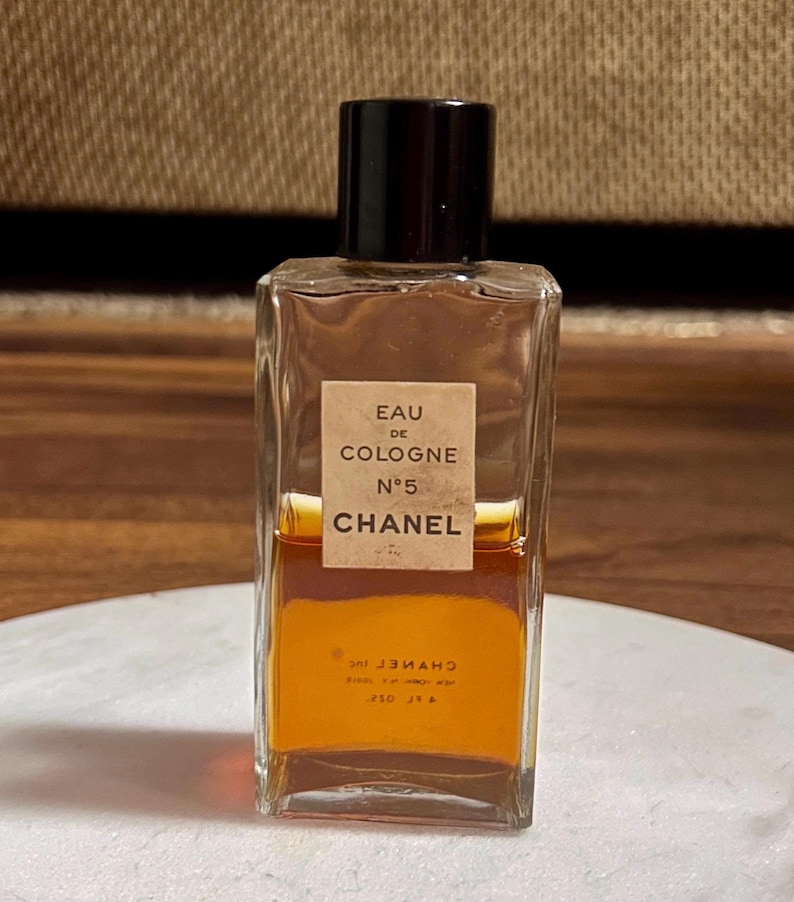 May include: A rectangular glass bottle of Chanel No. 5 Eau de Cologne. The bottle has a black cap and a beige label with black text. The liquid inside is amber-colored. The bottle is sitting on a white surface.