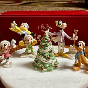 Może przedstawiać: Zestaw figurek bożonarodzeniowych Lenox z postaciami Disneya: Myszka Miki, Myszka Minnie, Kaczor Donald, Goofy i Pluto. Figurki znajdują się na białej podstawie z zieloną choinką pośrodku. Zestaw znajduje się przed czerwonym pudełkiem.