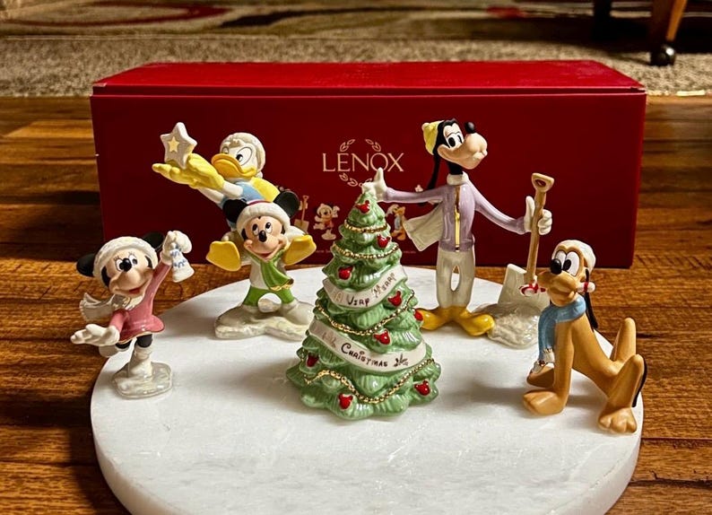 Może przedstawiać: Zestaw figurek bożonarodzeniowych Lenox z postaciami Disneya: Myszka Miki, Myszka Minnie, Kaczor Donald, Goofy i Pluto. Figurki znajdują się na białej podstawie z zieloną choinką pośrodku. Zestaw znajduje się przed czerwonym pudełkiem.