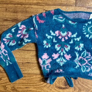 May include: A teal blue fuzzy sweater with a pink, green, and white floral pattern. The sweater has a loose fit and a slightly cropped length.