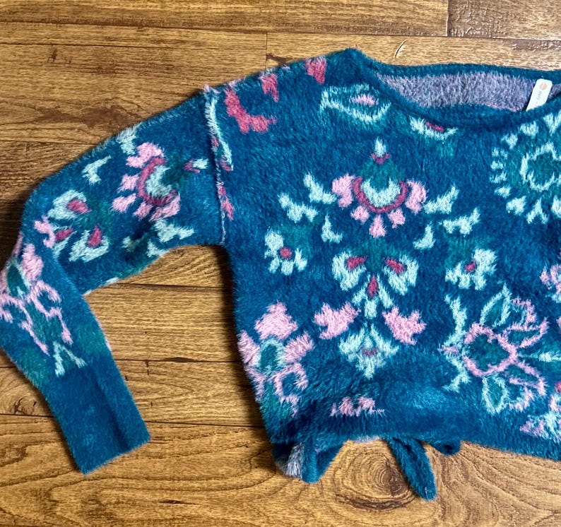 May include: A teal blue fuzzy sweater with a pink, green, and white floral pattern. The sweater has a loose fit and a slightly cropped length.