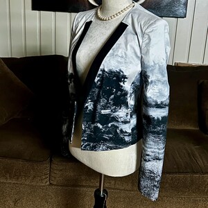 May include: A landscape-print blazer with black trim and a black and white forest design. The open-front jacket is displayed on a mannequin with a pearl necklace. The blazer's sleeves and body feature a scenic pattern.