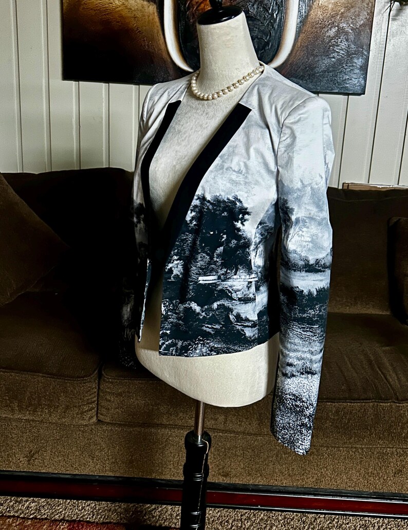 May include: A landscape-print blazer with black trim and a black and white forest design. The open-front jacket is displayed on a mannequin with a pearl necklace. The blazer's sleeves and body feature a scenic pattern.