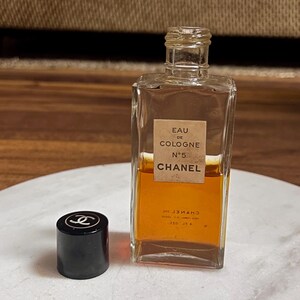 May include: A vintage Chanel No. 5 Eau de Cologne bottle with a black cap. The rectangular glass bottle contains amber-colored liquid and features a label with the brand name and product details. The cap has the Chanel logo.