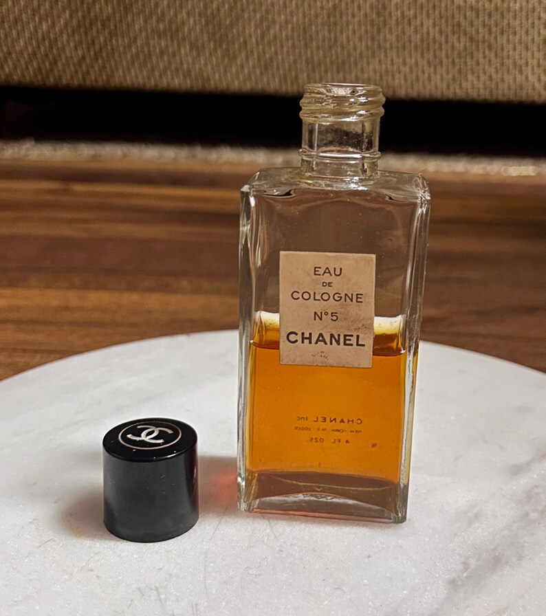 May include: A vintage Chanel No. 5 Eau de Cologne bottle with a black cap. The rectangular glass bottle contains amber-colored liquid and features a label with the brand name and product details. The cap has the Chanel logo.