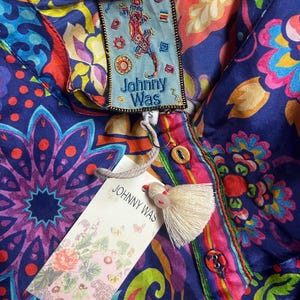 May include: A vibrant, patterned blouse with a button-down front and a tag that reads "Johnny Was." The blouse features a colorful floral and paisley design in shades of blue, red, pink, and green. A cream-colored tassel hangs from the tag.