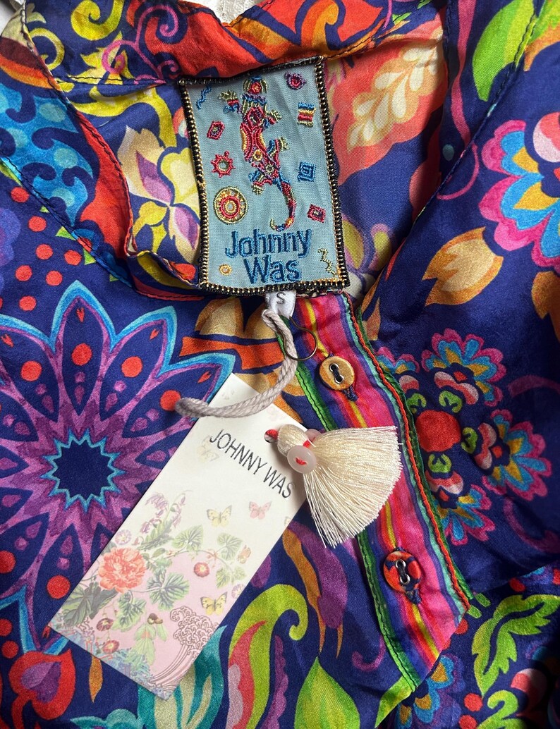 May include: A vibrant, patterned blouse with a button-down front and a tag that reads "Johnny Was." The blouse features a colorful floral and paisley design in shades of blue, red, pink, and green. A cream-colored tassel hangs from the tag.