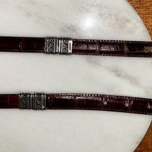 May include: Two brown leather belts with silver-toned metal accents. The belts have a textured, crocodile-like pattern. One belt has a buckle with a yellow tag. The belts are displayed on a white marble surface.
