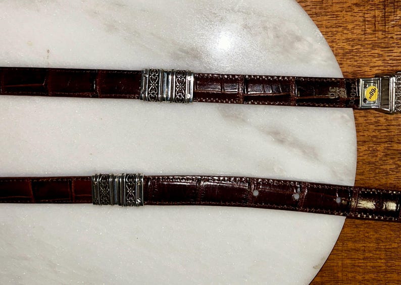 May include: Two brown leather belts with silver-toned metal accents. The belts have a textured, crocodile-like pattern. One belt has a buckle with a yellow tag. The belts are displayed on a white marble surface.