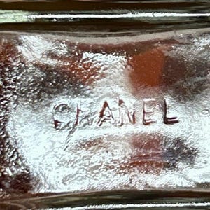 May include: Close-up of the bottom of a clear glass bottle with the embossed word "CHANEL". The glass has a textured surface and reflects light, creating highlights and shadows. The overall aesthetic is simple and elegant.