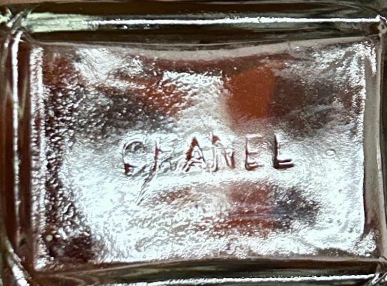 May include: Close-up of the bottom of a clear glass bottle with the embossed word "CHANEL". The glass has a textured surface and reflects light, creating highlights and shadows. The overall aesthetic is simple and elegant.
