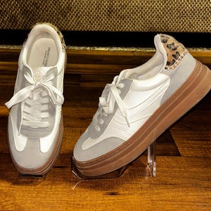 May include: A pair of white and gray sneakers with brown soles. The sneakers have white laces and a leopard print detail on the heel. The shoes are displayed on clear stands against a wooden background.
