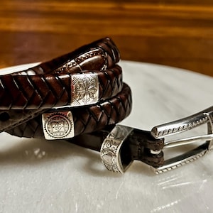 K&ouml;nnte beinhalten: Drei braune Lederg&uuml;rtel mit silbernen Schnallen. Die G&uuml;rtel sind geflochten und haben dekorative silberne Akzente. Die Schnallen haben aufwendige Designs. Die G&uuml;rtel werden auf einer wei&szlig;en Oberfl&auml;che pr&auml;sentiert.