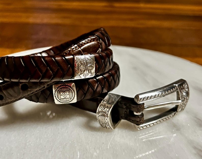 K&ouml;nnte beinhalten: Drei braune Lederg&uuml;rtel mit silbernen Schnallen. Die G&uuml;rtel sind geflochten und haben dekorative silberne Akzente. Die Schnallen haben aufwendige Designs. Die G&uuml;rtel werden auf einer wei&szlig;en Oberfl&auml;che pr&auml;sentiert.