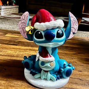 May include: A blue Stitch figurine wearing a Santa hat and a small bell. A smaller Stitch figure is also present, wearing a Santa hat. The figurine is on a white base, with a Christmas theme.