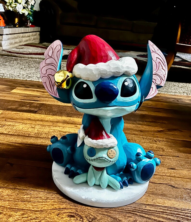 May include: A blue Stitch figurine wearing a Santa hat and a small bell. A smaller Stitch figure is also present, wearing a Santa hat. The figurine is on a white base, with a Christmas theme.