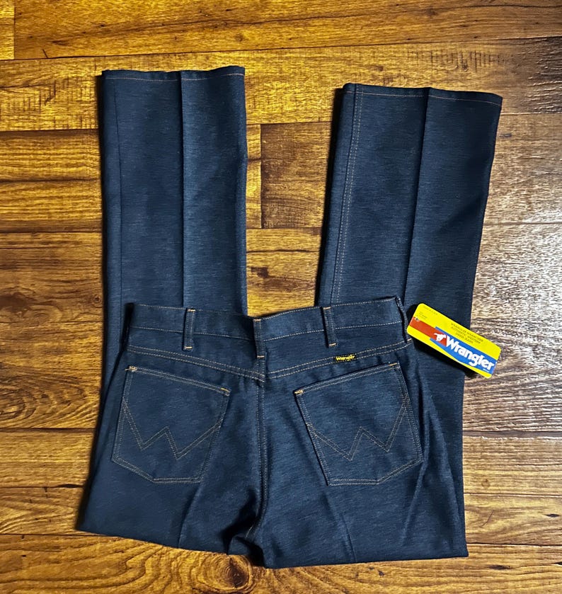 Może przedstawiać: Ciemnoniebieskie dżinsy Wrangler z klasycznym pięciokieszeniowym designem. Dżinsy mają prosty kr&oacute;j nogawek i logo Wrangler na tylnej kieszeni oraz ż&oacute;łto-czerwoną metkę. Dżinsy leżą płasko na drewnianej powierzchni.