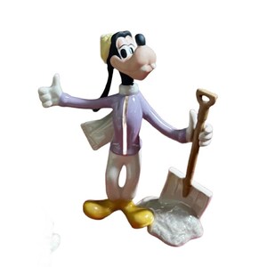 Może przedstawiać: Figurka Goofy'ego, postaci z kresk&oacute;wek Disneya, w jasnofioletowej kurtce, białych spodniach i ż&oacute;łtym kapeluszu. Trzyma łopatę i pokazuje kciuk w g&oacute;rę. Figurka stoi na białej podstawie.