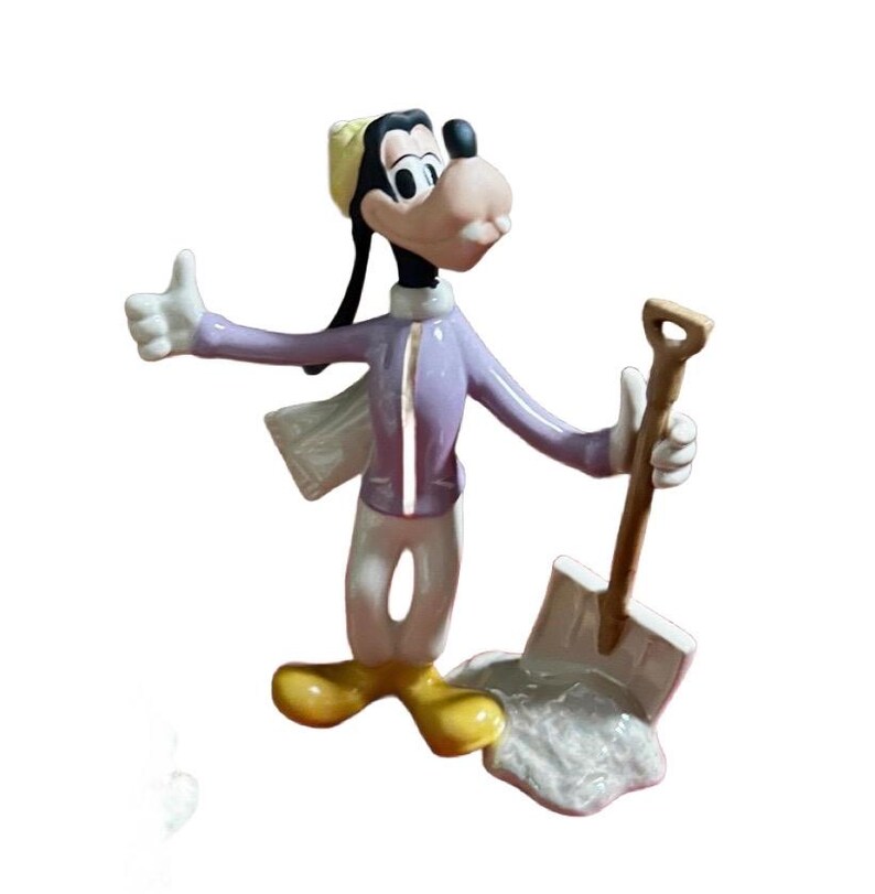 Może przedstawiać: Figurka Goofy'ego, postaci z kresk&oacute;wek Disneya, w jasnofioletowej kurtce, białych spodniach i ż&oacute;łtym kapeluszu. Trzyma łopatę i pokazuje kciuk w g&oacute;rę. Figurka stoi na białej podstawie.