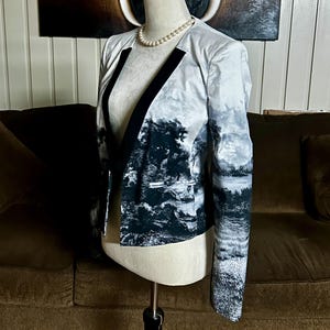 May include: A blazer with a landscape print in shades of gray and black. The jacket has a black lapel and a classic cut. The sleeves are long, and the jacket is displayed on a mannequin.