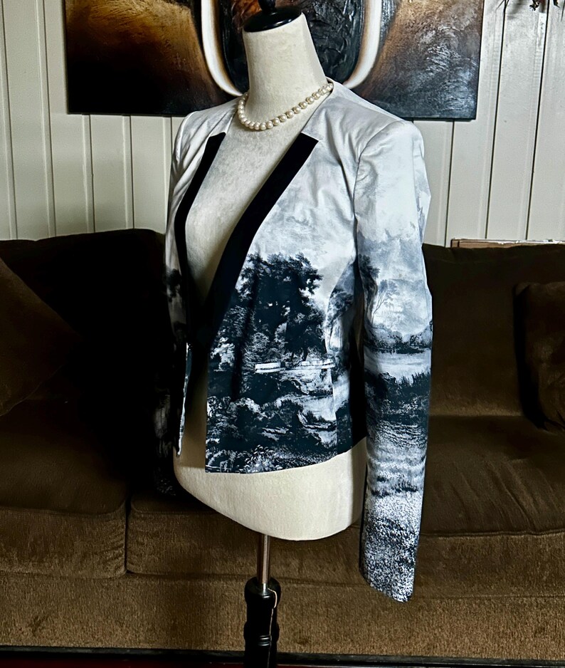 May include: A blazer with a landscape print in shades of gray and black. The jacket has a black lapel and a classic cut. The sleeves are long, and the jacket is displayed on a mannequin.
