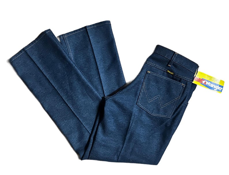 Może przedstawiać: Ciemnoniebieskie dżinsy Wrangler z denimu o klasycznym kroju. Dżinsy mają konstrukcję tylnej kieszeni z białymi przeszyciami oraz ż&oacute;łtą i białą metkę Wrangler. Dżinsy są złożone i ułożone płasko.