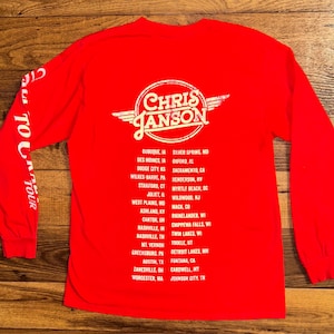 May include: Red long-sleeve shirt featuring the text "CHRIS JANSON" in a white, circular design with wings. The back of the shirt lists tour locations in white text. The left sleeve has the text "TOUR" vertically.