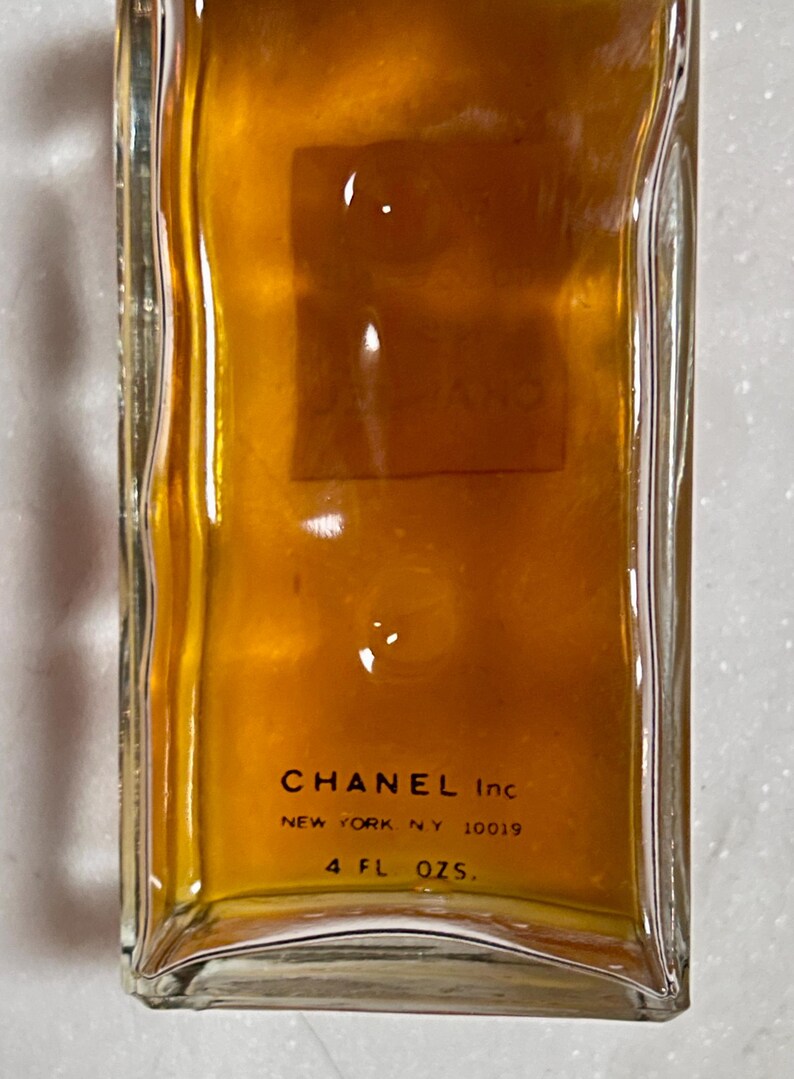 May include: A rectangular glass bottle filled with a golden liquid. The bottom of the bottle features the text "CHANEL Inc NEW YORK, N.Y. 10019" and "4 FL. OZS."