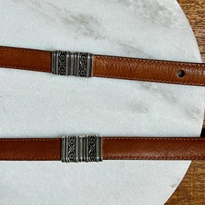 May include: Two brown leather belts with silver-colored decorative clasps. The belts are displayed on a white marble surface. The clasps have an ornate design.