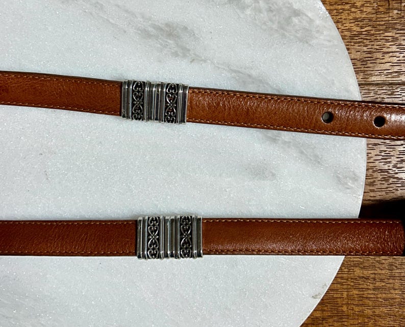 May include: Two brown leather belts with silver-colored decorative clasps. The belts are displayed on a white marble surface. The clasps have an ornate design.