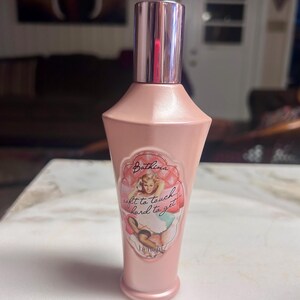 May include: A pink cosmetic bottle with a silver cap. The bottle has a label with a vintage pin-up illustration and the text "Bathina soft to touch... hard to get". The product is from the brand Benefit.