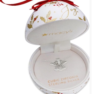 May include: A white Christmas ornament with gold holly and berry accents, and the word "Joy" in gold script. Inside, a silver angel pendant necklace with cubic zirconia is displayed on a white velvet backing. The box reads "Cubic Zirconia Sterling Silver."