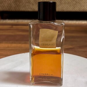 May include: A rectangular Chanel cologne bottle with a black cap. The clear glass bottle contains a golden-brown liquid. The label reads "CHANEL Inc." and "COLOGNE N&deg;5". The bottle is sitting on a white surface.