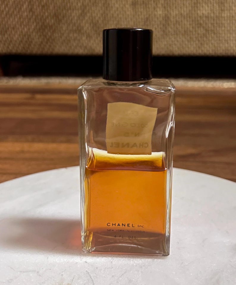 May include: A rectangular Chanel cologne bottle with a black cap. The clear glass bottle contains a golden-brown liquid. The label reads "CHANEL Inc." and "COLOGNE N&deg;5". The bottle is sitting on a white surface.
