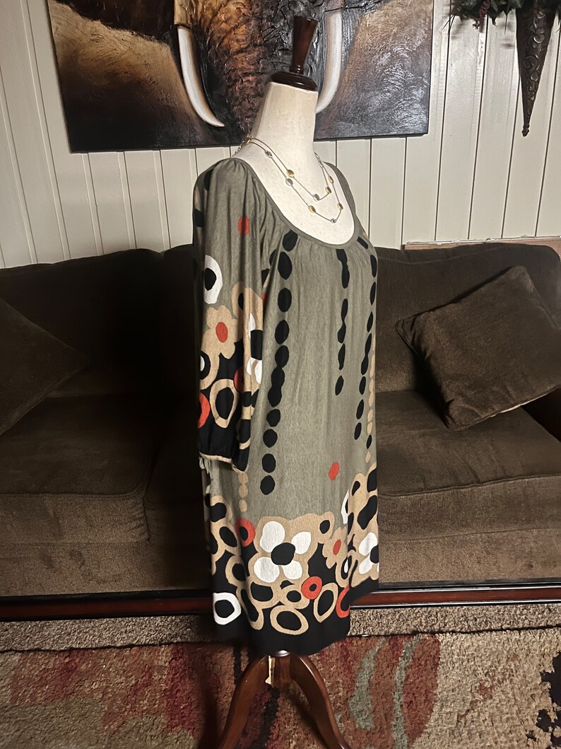 May include: A green, black, white, and orange patterned dress with 3/4 sleeves. The dress is on a white mannequin with a gold and silver necklace.