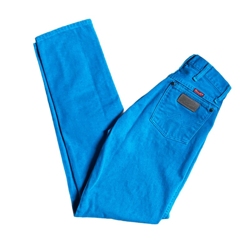K&ouml;nnte beinhalten: Helle blaue Wrangler-Jeans. Die Jeans haben ein klassisches F&uuml;nf-Taschen-Design, einen braunen Lederaufn&auml;her auf der Ges&auml;&szlig;tasche und das Wrangler-Logo. Die Jeans sind auf wei&szlig;em Hintergrund gefaltet.