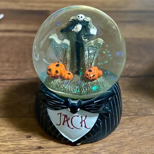 Puede incluir: Una bola de nieve de Jack Skellington con Jack frente a l&aacute;pidas y calabazas naranjas en la base. La base es negra con un emblema blanco en forma de coraz&oacute;n que dice "Jack". El globo est&aacute; lleno de purpurina.
