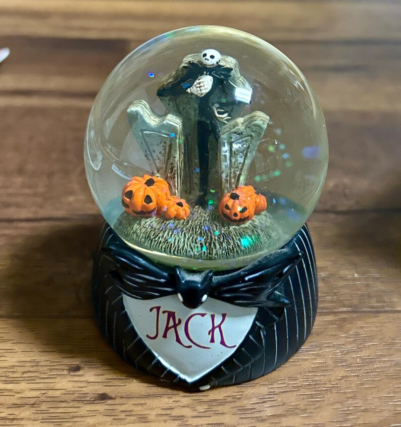Puede incluir: Una bola de nieve de Jack Skellington con Jack frente a l&aacute;pidas y calabazas naranjas en la base. La base es negra con un emblema blanco en forma de coraz&oacute;n que dice "Jack". El globo est&aacute; lleno de purpurina.