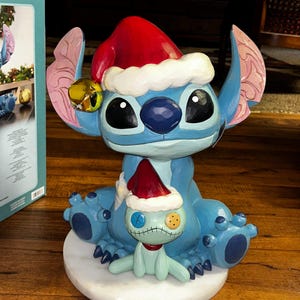 May include: A blue Stitch figurine wearing a red Santa hat with white trim and a gold bell. A smaller Stitch figure in a Santa hat sits on a white base. The figurine is a Christmas decoration.