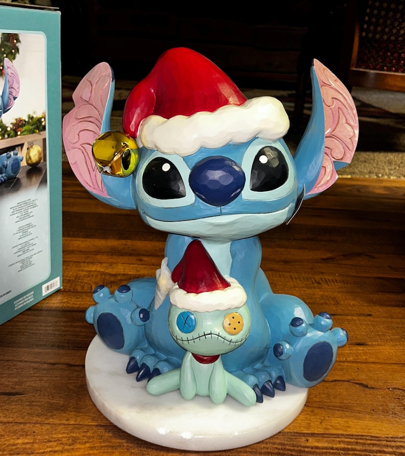 May include: A blue Stitch figurine wearing a red Santa hat with white trim and a gold bell. A smaller Stitch figure in a Santa hat sits on a white base. The figurine is a Christmas decoration.
