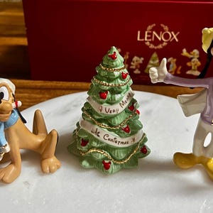 Może przedstawiać: Kolekcja figurek świątecznych Lenox z postaciami Disneya. Figurki obejmują Plutona, Goofy'ego i choinkę z banerem z napisem "Very Merry Christmas". Drzewo jest zielone z czerwonymi ozdobami.