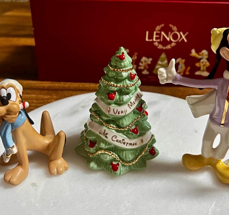 Może przedstawiać: Kolekcja figurek świątecznych Lenox z postaciami Disneya. Figurki obejmują Plutona, Goofy'ego i choinkę z banerem z napisem "Very Merry Christmas". Drzewo jest zielone z czerwonymi ozdobami.