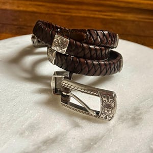 K&ouml;nnte beinhalten: Geflochtener brauner Lederg&uuml;rtel mit silberner Schnalle. Der G&uuml;rtel besteht aus gewebtem Leder und verf&uuml;gt &uuml;ber eine dekorative Silberschnalle mit einem eingravierten Design. Der G&uuml;rtel wird aufgerollt und tragbar gezeigt.