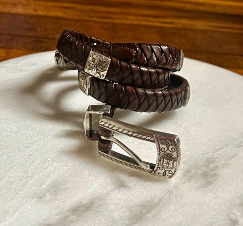 K&ouml;nnte beinhalten: Geflochtener brauner Lederg&uuml;rtel mit silberner Schnalle. Der G&uuml;rtel besteht aus gewebtem Leder und verf&uuml;gt &uuml;ber eine dekorative Silberschnalle mit einem eingravierten Design. Der G&uuml;rtel wird aufgerollt und tragbar gezeigt.